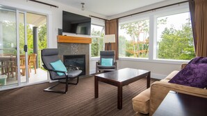 A modern living room with a fireplace, two armchairs, a coffee table, and large windows offering a view of the outdoors. - Ocean Tide (Unit A+B) - Waterfront Condo - Sleeps 6 - Pool - Family (Nanoose Bay)