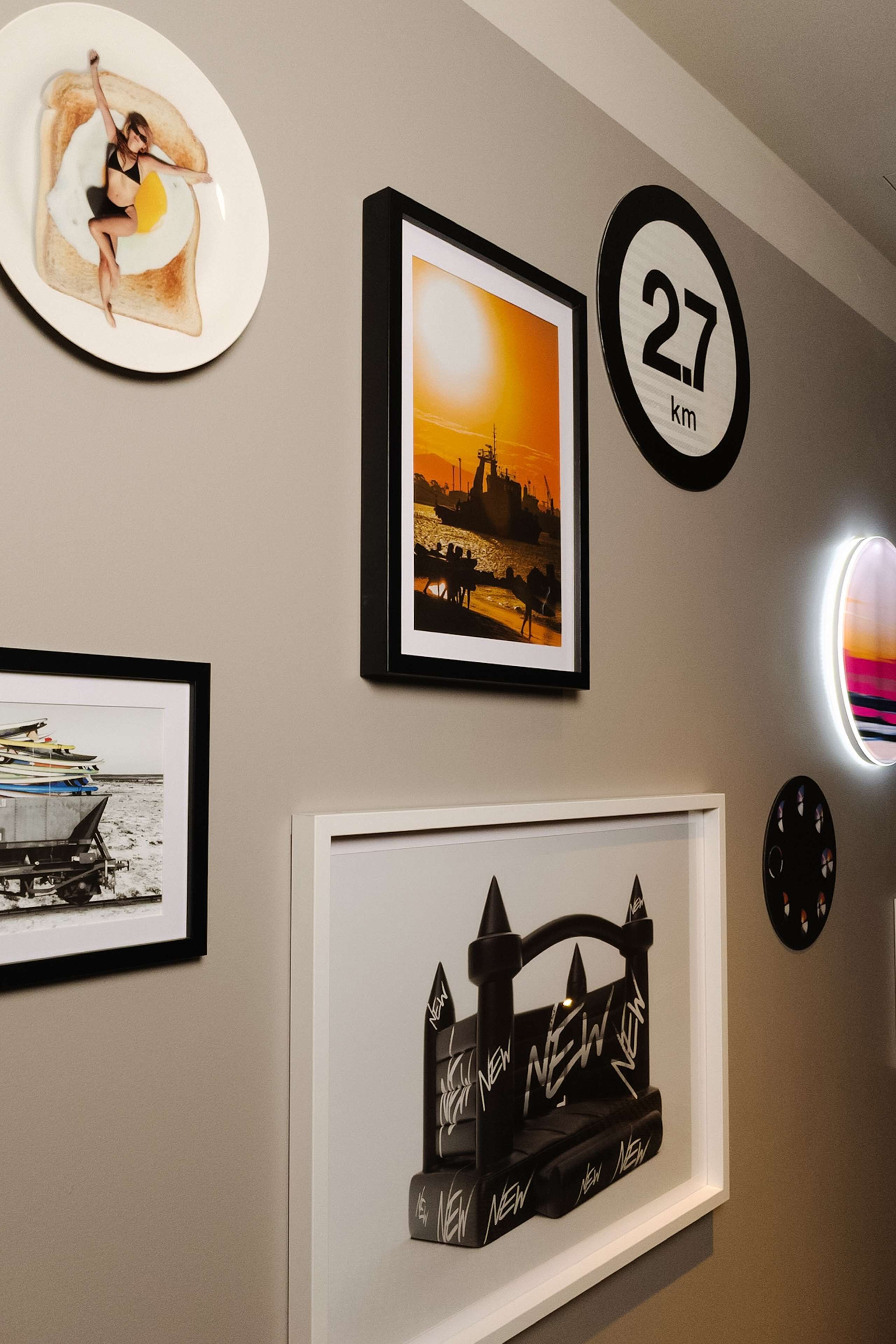 A wall with various framed artworks, including a sunset cityscape, a silhouette of a bridge, and a person on toast. A round sign indicates a distance of 27 km.