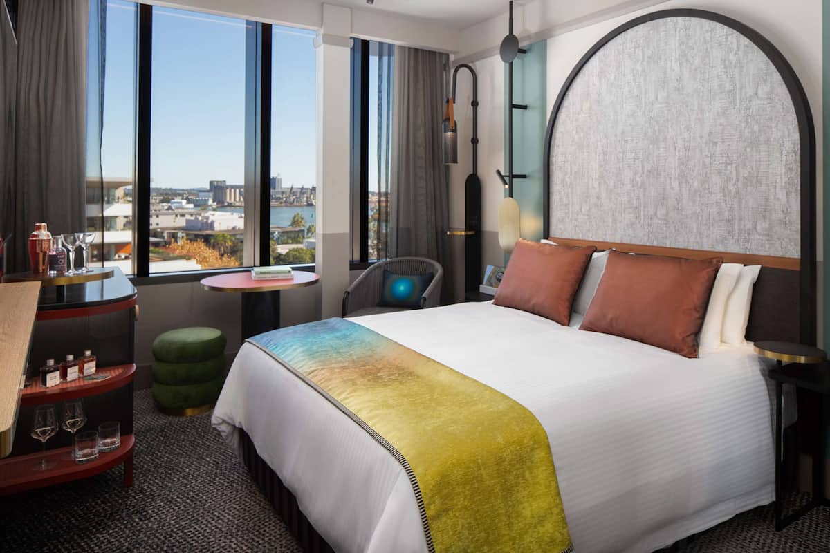 Una habitación de hotel moderna con una cama grande, vistas a la ciudad, una mesita con una silla y un reposapiés verde.