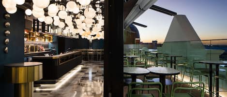 Zona de bar con piso de tablones, sillas verdes y luces esféricas colgantes.