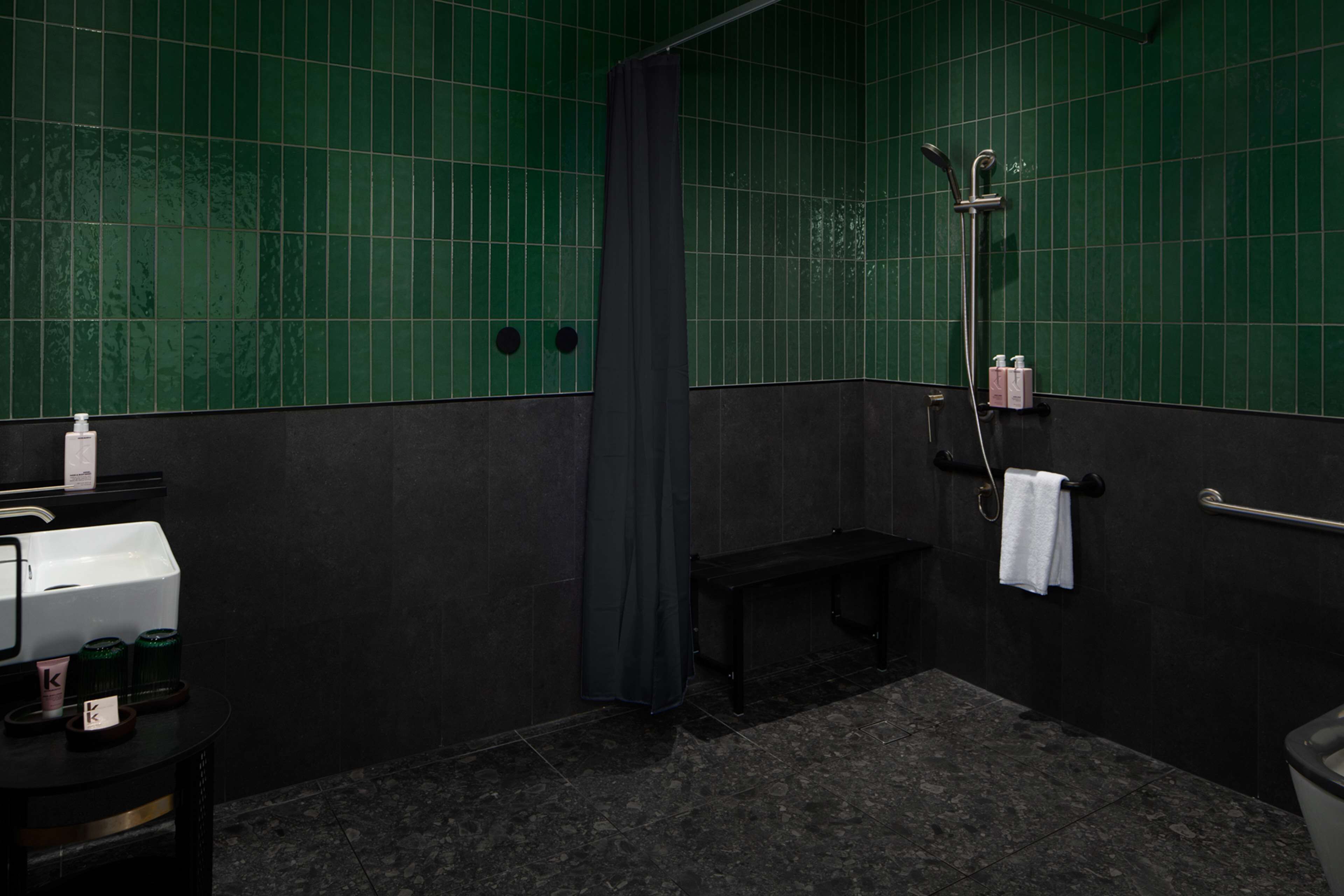 A bathroom with green tiled walls, a bench, a shower, and a sink with toiletries.