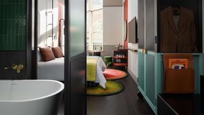 A modern bathroom with a freestanding bathtub, a bed with pillows, a small table with a chair, and a shelf with toiletries. - QT Newcastle (Newcastle)