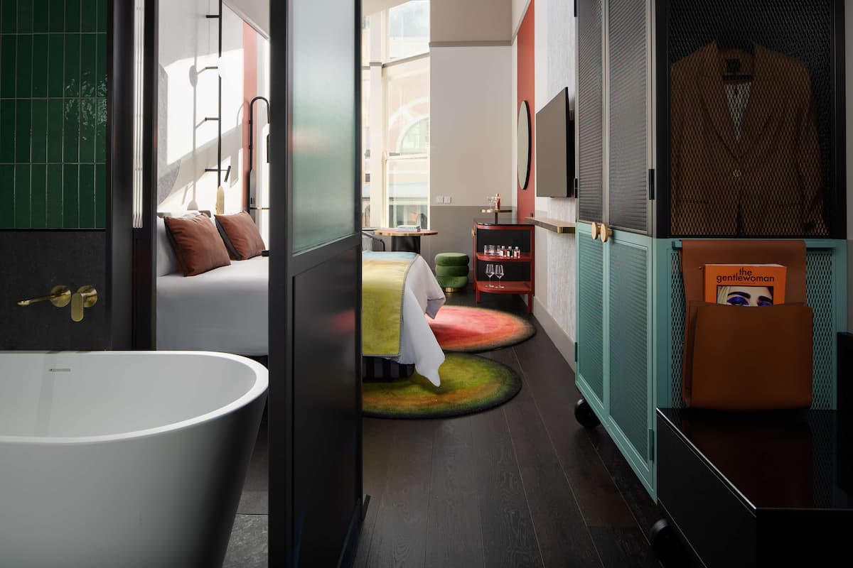 Un baño moderno con una bañera independiente, una cama con almohadas, una mesita con una silla y un estante con artículos de aseo.