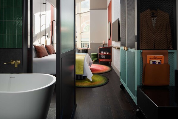 Un baño moderno con una bañera independiente, una cama con almohadas, una mesita con una silla y un estante con artículos de aseo.