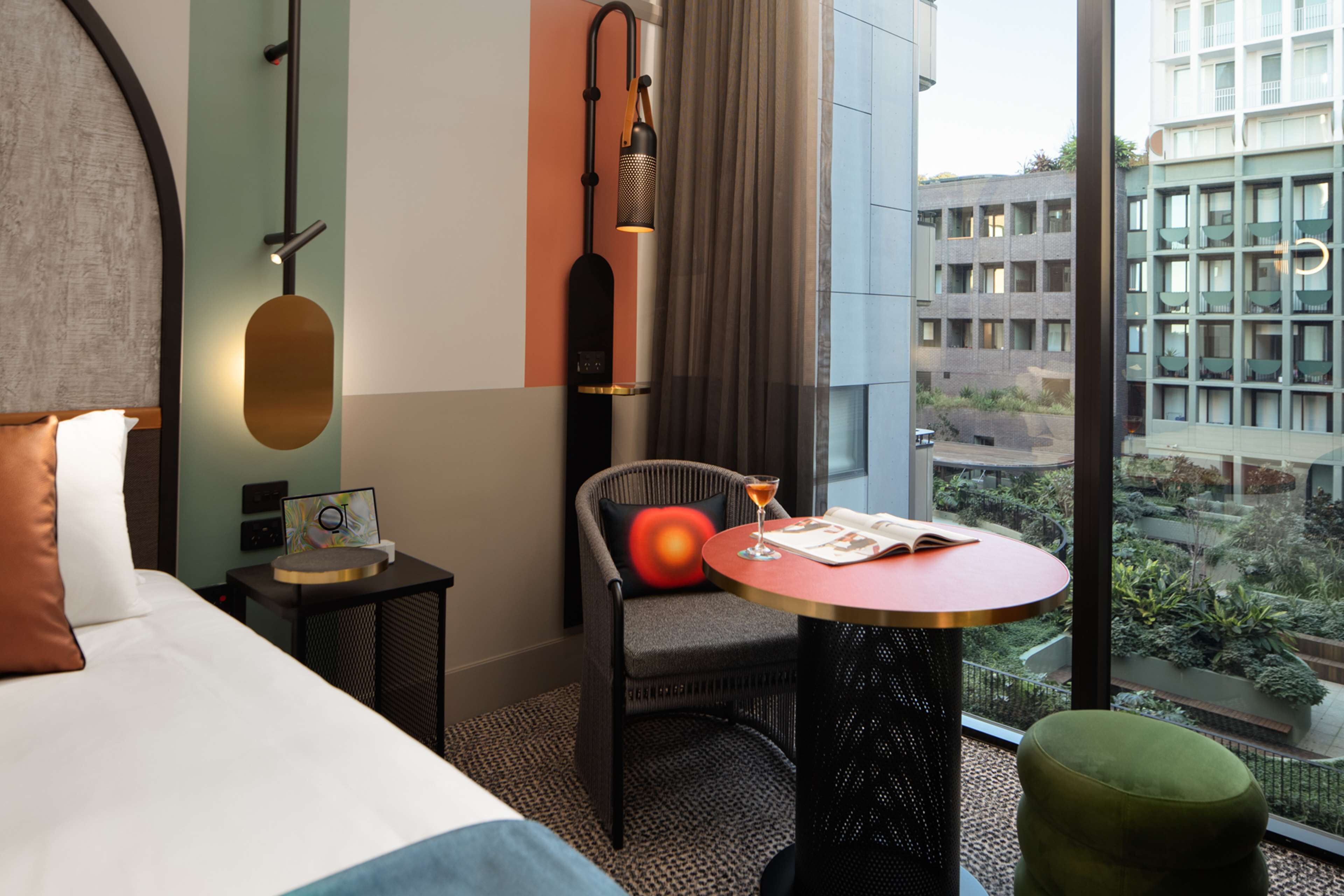 A hotel room with a bed, a round table with a drink, a chair, a small side table, and a view of the city through large windows.