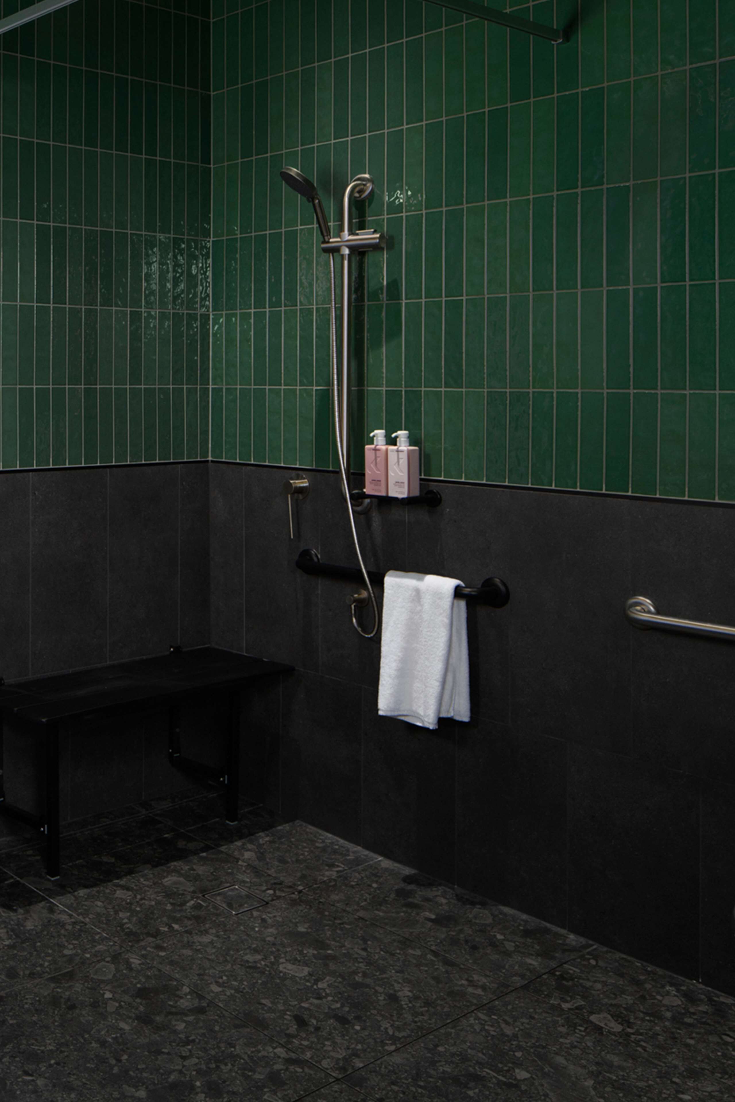 A bathroom with green tiled walls, a shower with a handheld showerhead, a bench, and a towel rack with a towel.