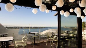 A restaurant with outdoor seating overlooking a body of water, featuring round tables and chairs. - QT Newcastle (Newcastle)