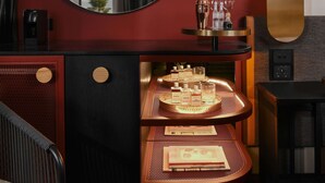 A modern hotel room with a red accent wall, a round mirror, a minibar, and a view of the cityscape through the window. - QT Newcastle (Newcastle)