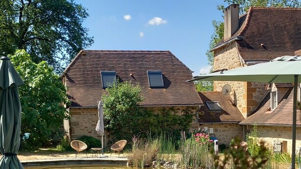 A wooden deck with lounge chairs, a small pond, stone buildings with tiled roofs, and a clear blue sky. - Quercy cottage, charm, comfort, garden with trees, natural swimming pool (Frontenac)