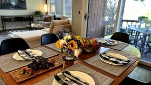 Un coin repas avec une table en bois dressée pour quatre personnes, agrémentée de sets de table, d’assiettes, de couverts et d’un centre de table composé de tournesols et de fruits.