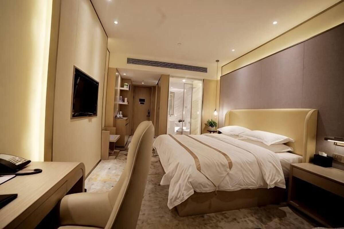 Quarto de hotel com cama, escrivaninha, cadeira, televisão e banheiro visível por uma porta aberta.