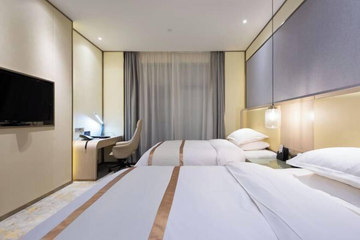 Quarto de hotel com cama, escrivaninha, cadeira, televisão e janela com cortinas.