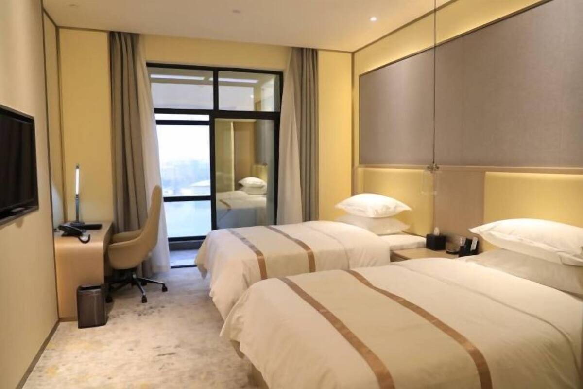 Quarto de hotel com duas camas, uma escrivaninha, uma cadeira, uma TV e uma janela grande.
