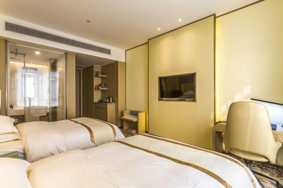 Habitación de hotel con dos camas, un escritorio, una silla, un televisor y un baño visible a través de una puerta de cristal.