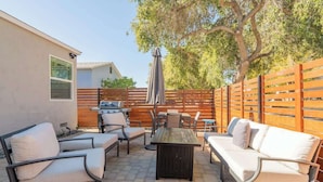 Une terrasse extérieure avec une table en bois, des chaises blanches avec coussins, un barbecue et une clôture en bois.