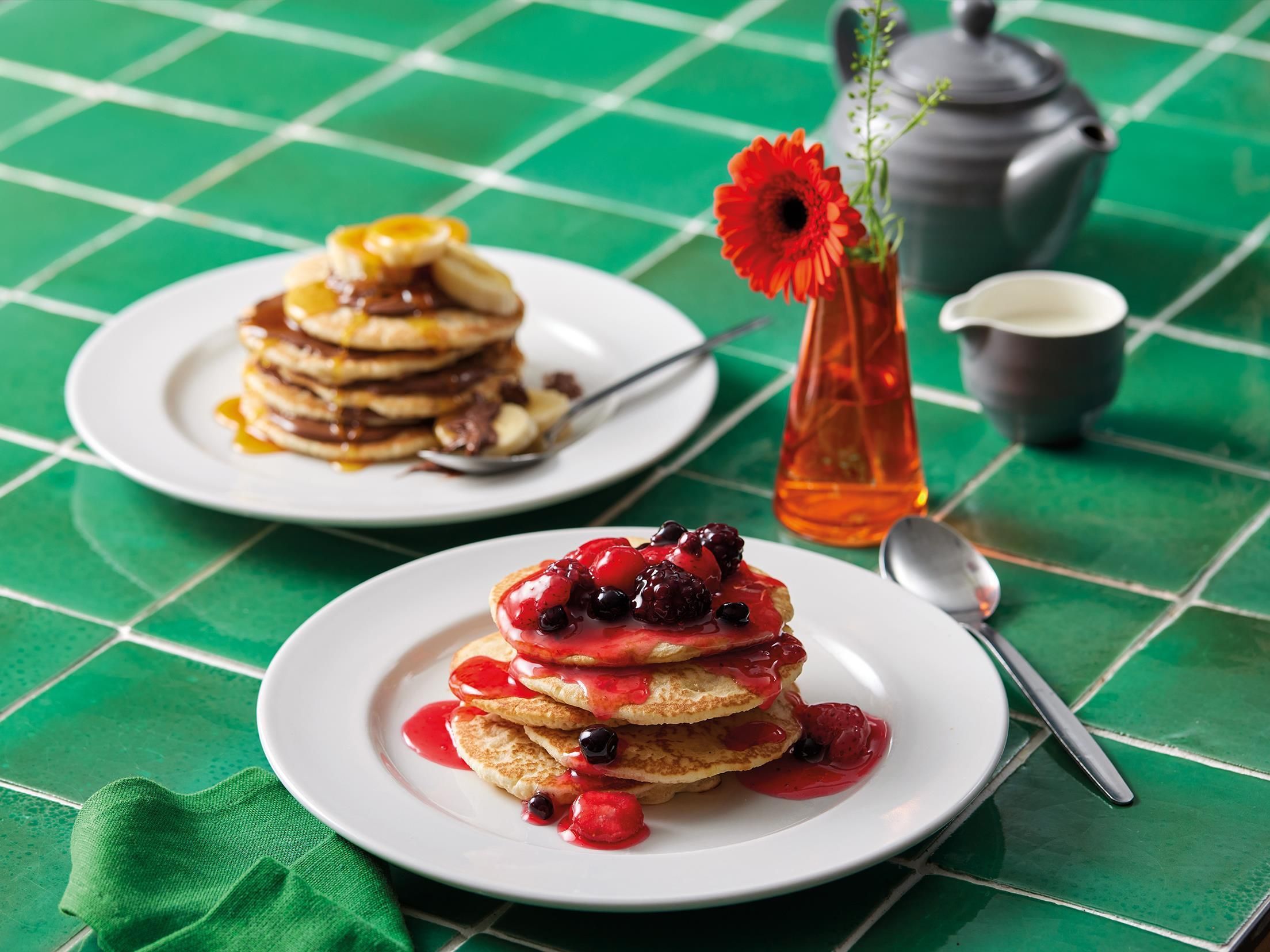 Ein Frühstückstisch mit Pfannkuchen und Beeren auf Tellern, einer Vase mit einer Blume und einem Teekännchen auf einer grün karierten Tischdecke.