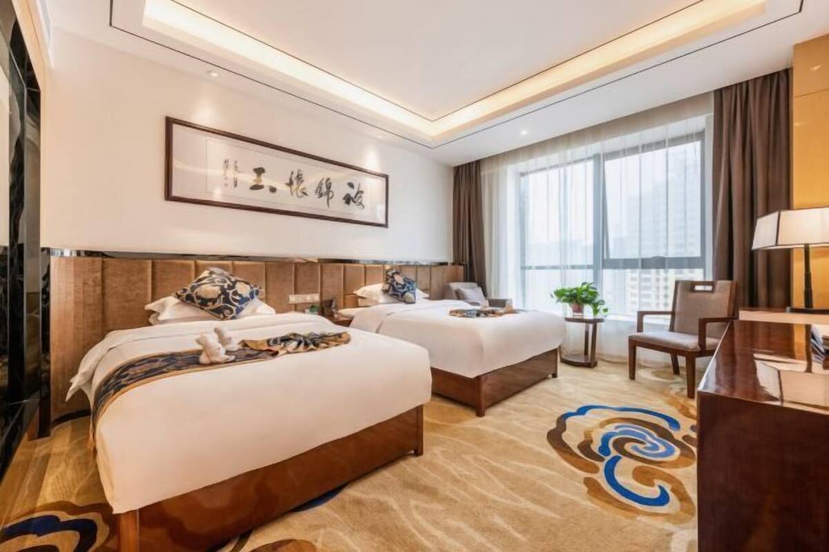 A hotel room with two beds, a desk, a chair, a window with curtains, and a framed artwork on the wall.