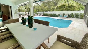 Un espace aménagé au bord de la piscine, avec une table et des chaises, un ventilateur de plafond, et une vue sur la piscine et la verdure environnante.