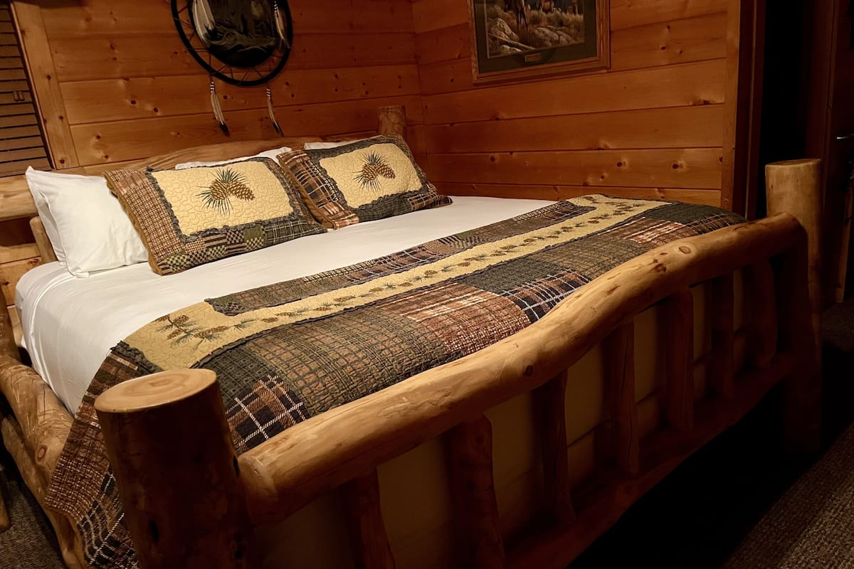 A wooden bed with a quilt and pillows, a wooden headboard, and a framed picture on the wall.