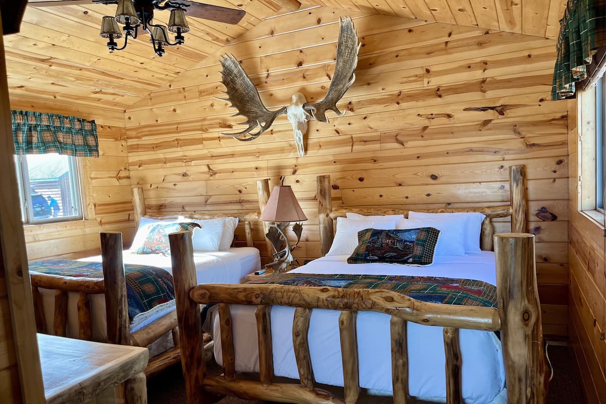 A wooden cabin bedroom with two beds, a chandelier, and a mounted deer head.