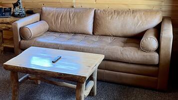 Un sofá de cuero color bronceado con una mesa de centro de madera, una lámpara y un teléfono sobre una mesita auxiliar de madera.