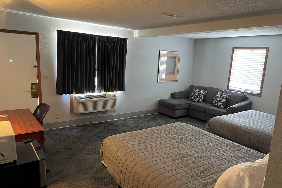 A hotel room with a desk, two beds, a sofa, a window with blinds, an air conditioning unit, and a framed picture on the wall.