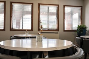A dining area with round tables and chairs, large windows with blinds, and a potted plant.