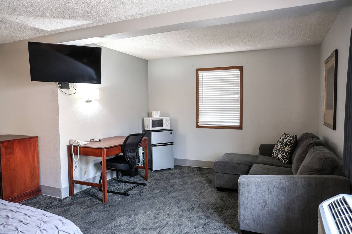 A compact room with a bed, a desk with a chair, a kitchenette, a gray sofa, and a television mounted on the wall.