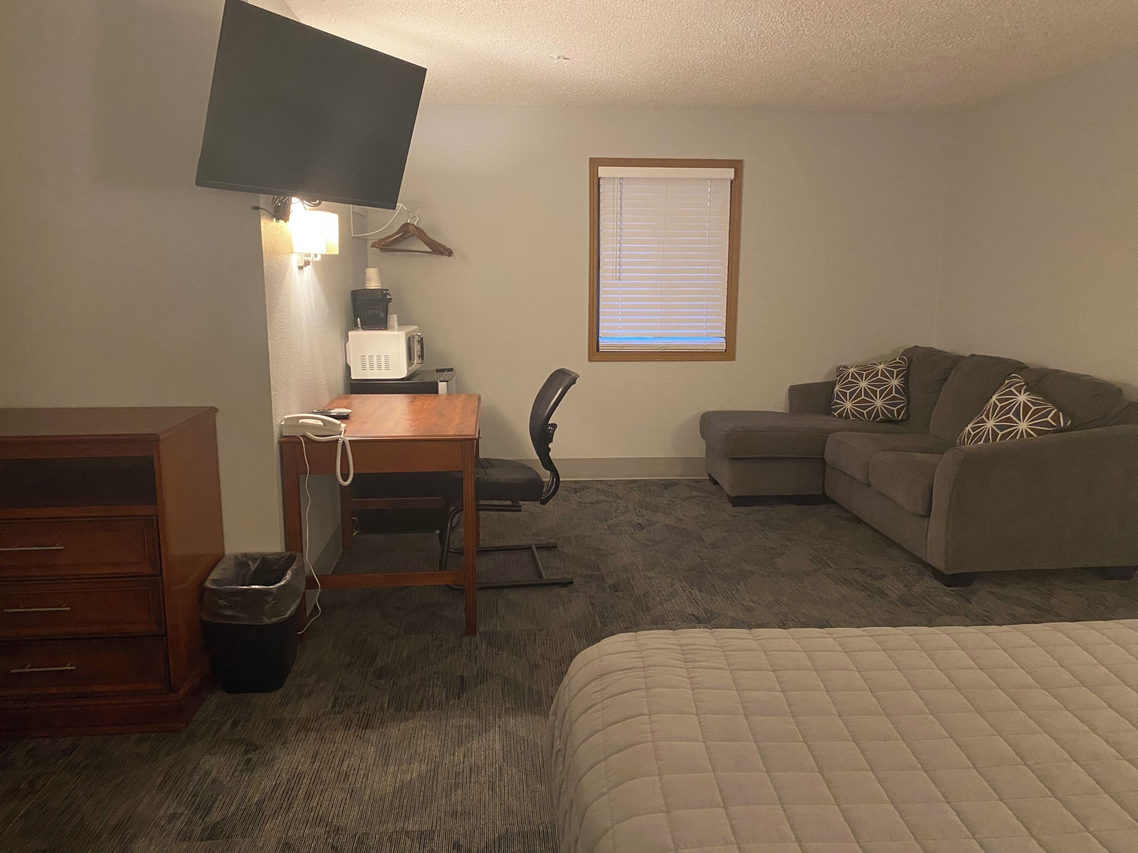 A hotel room with a bed, a desk with a telephone, a chair, a sofa, a television mounted on the wall, and a window with blinds.