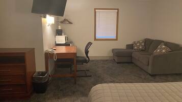 A hotel room with a bed, a desk with a telephone, a chair, a sofa, a television mounted on the wall, and a window with blinds.