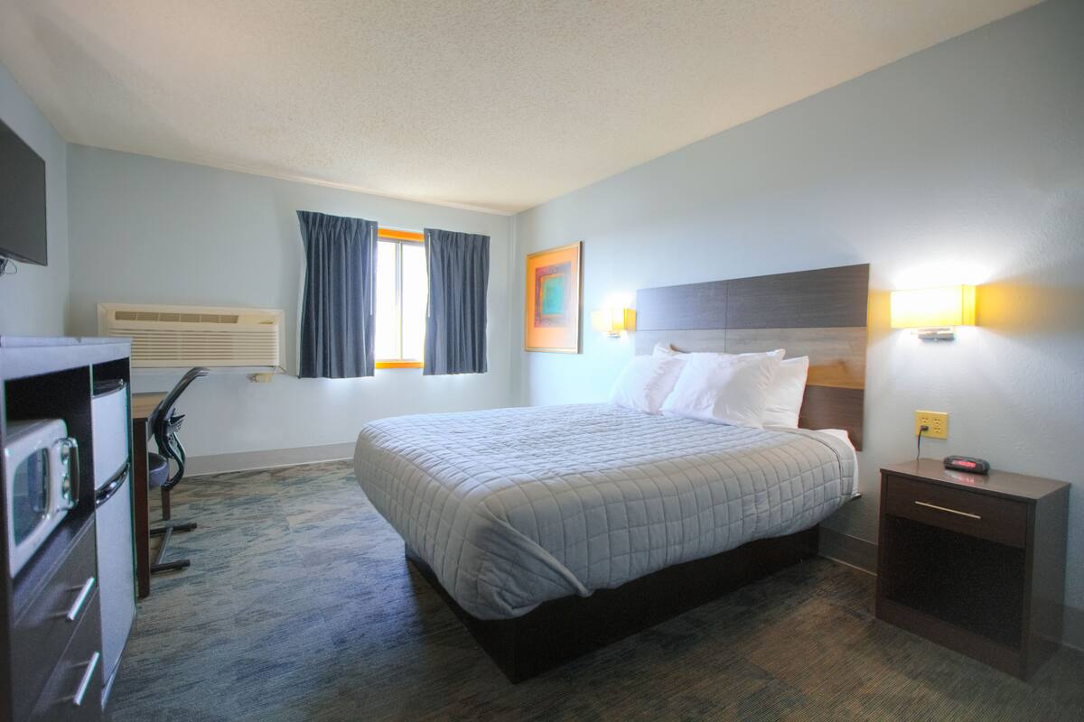 A hotel room with a bed, a desk, a chair, a dresser, a window with curtains, and a nightstand with a lamp.