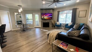 Een ruime woonkamer met een open haard, een plafondventilator en een aan de muur bevestigde televisie.