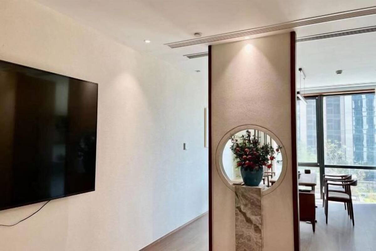 Et moderne interiør med et stort fladskærms-tv på væggen, et rundt spejl med en vase blomster, og udsigt til et spiseområde gennem et stort vindue.