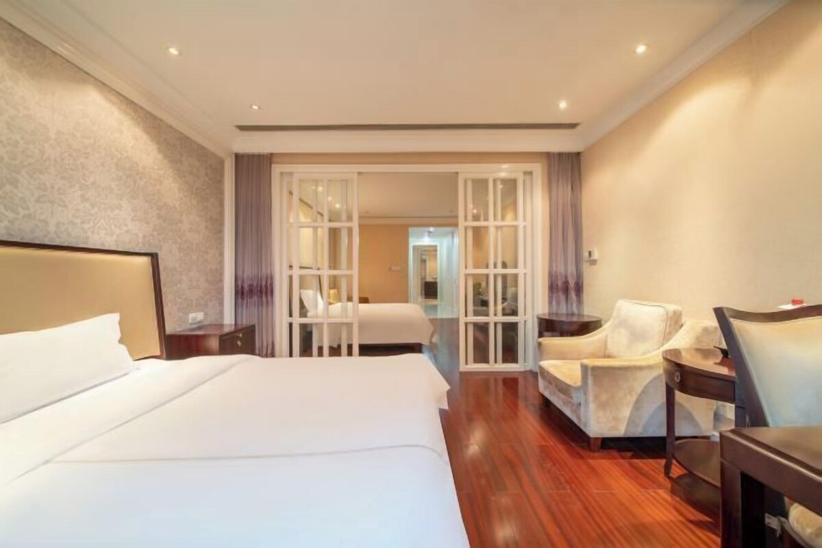 A hotel room with a large bed, a sitting area with a chair, a small table, and a view through a glass door.