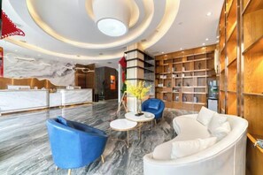 Un hall de hotel con mostrador de recepción, zona de estar con sofás y sillones, suelo de mármol y una estantería.