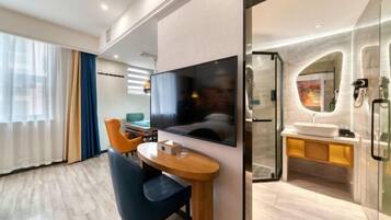 A modern hotel room with a wooden dining table, a flat-screen TV, a glass-enclosed bathroom with a sink and mirror, and a view of the city through large windows.