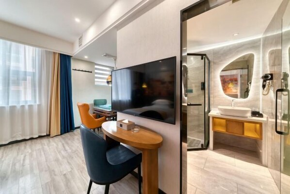 A modern hotel room with a wooden dining table, a flat-screen TV, a glass-enclosed bathroom with a sink and mirror, and a view of the city through large windows.