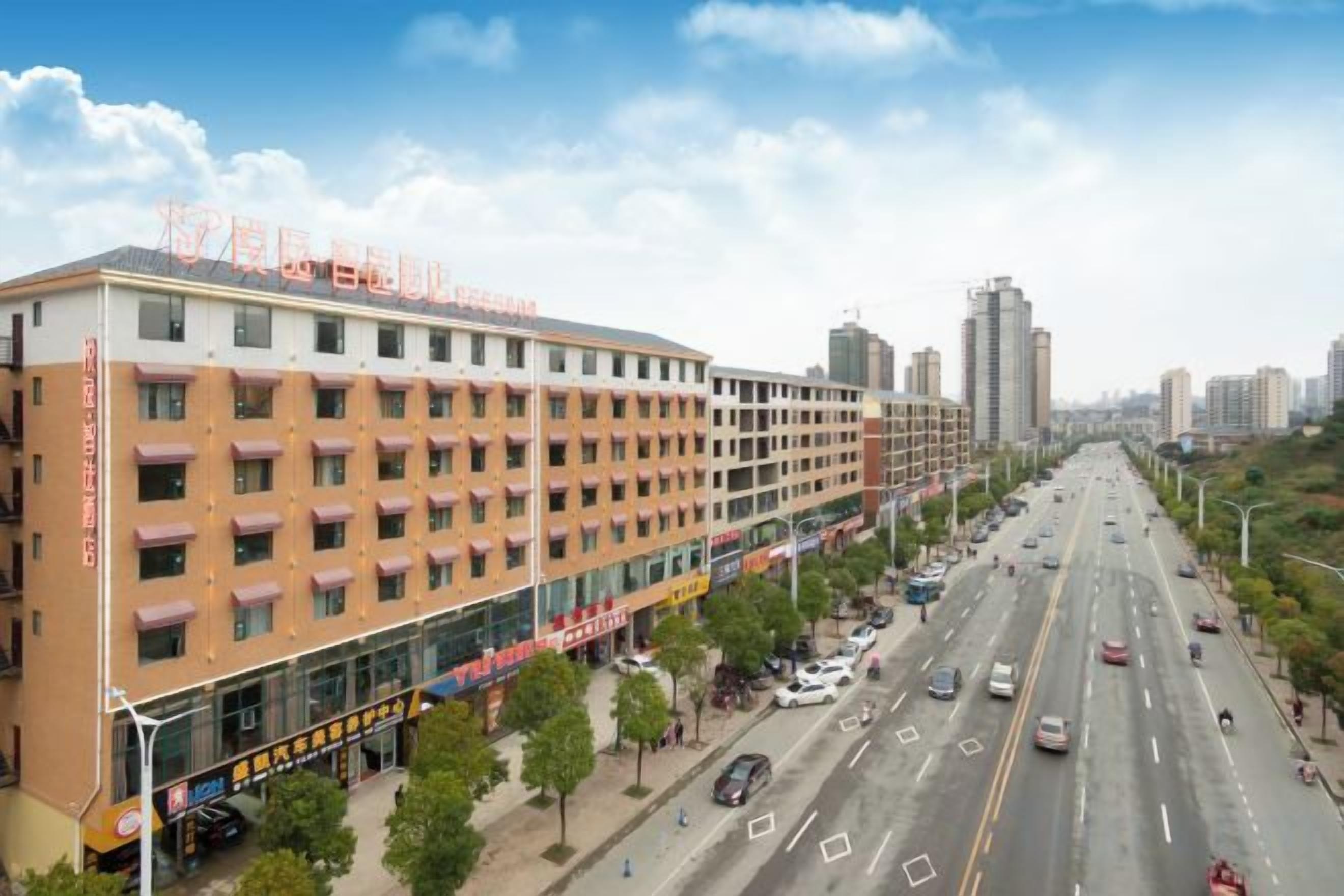 A multi-story hotel with a row of rooms on the ground floor, a parking lot, and a street view with vehicles and trees.
