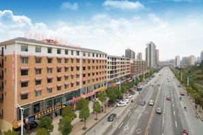 Et hotel med flere etager, en række værelser i stueplan, en parkeringsplads og udsigt til gaden med biler og træer.