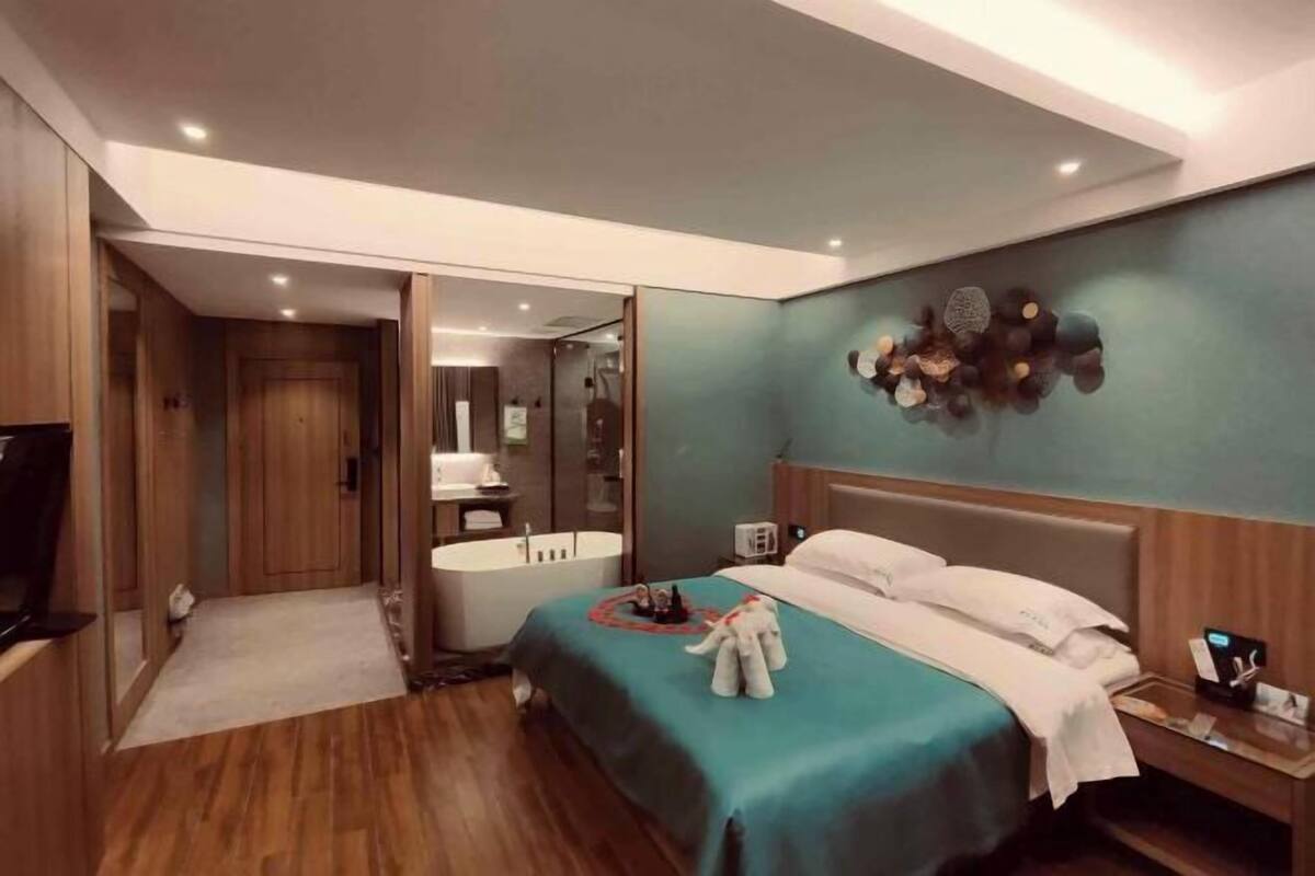 Une chambre d’hôtel avec un lit, une table de chevet, une horloge fixée au mur et une salle de bain visible à travers une porte ouverte.