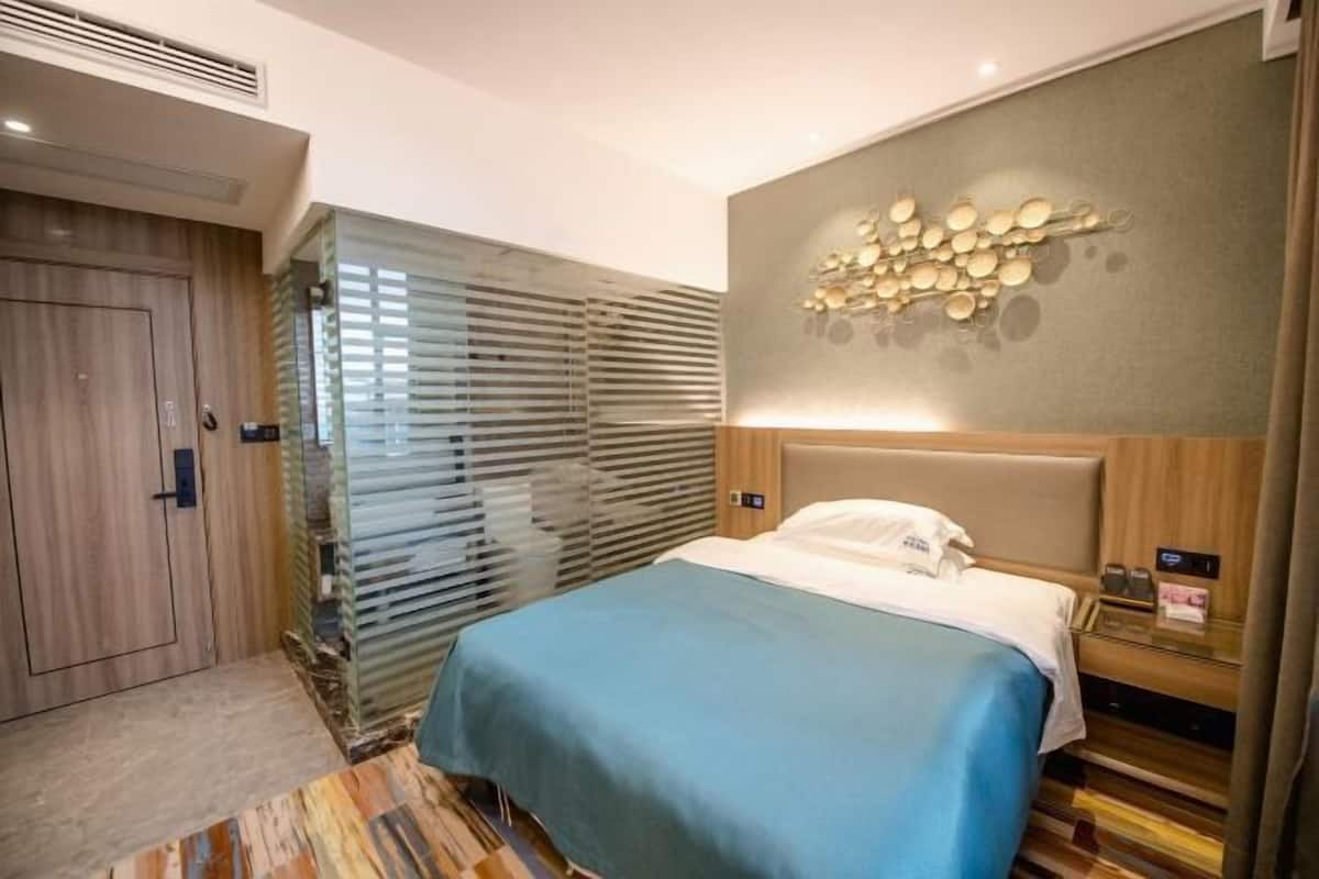 Une chambre d’hôtel avec un lit, un luminaire fixé au mur, une porte en bois et une fenêtre avec des stores.