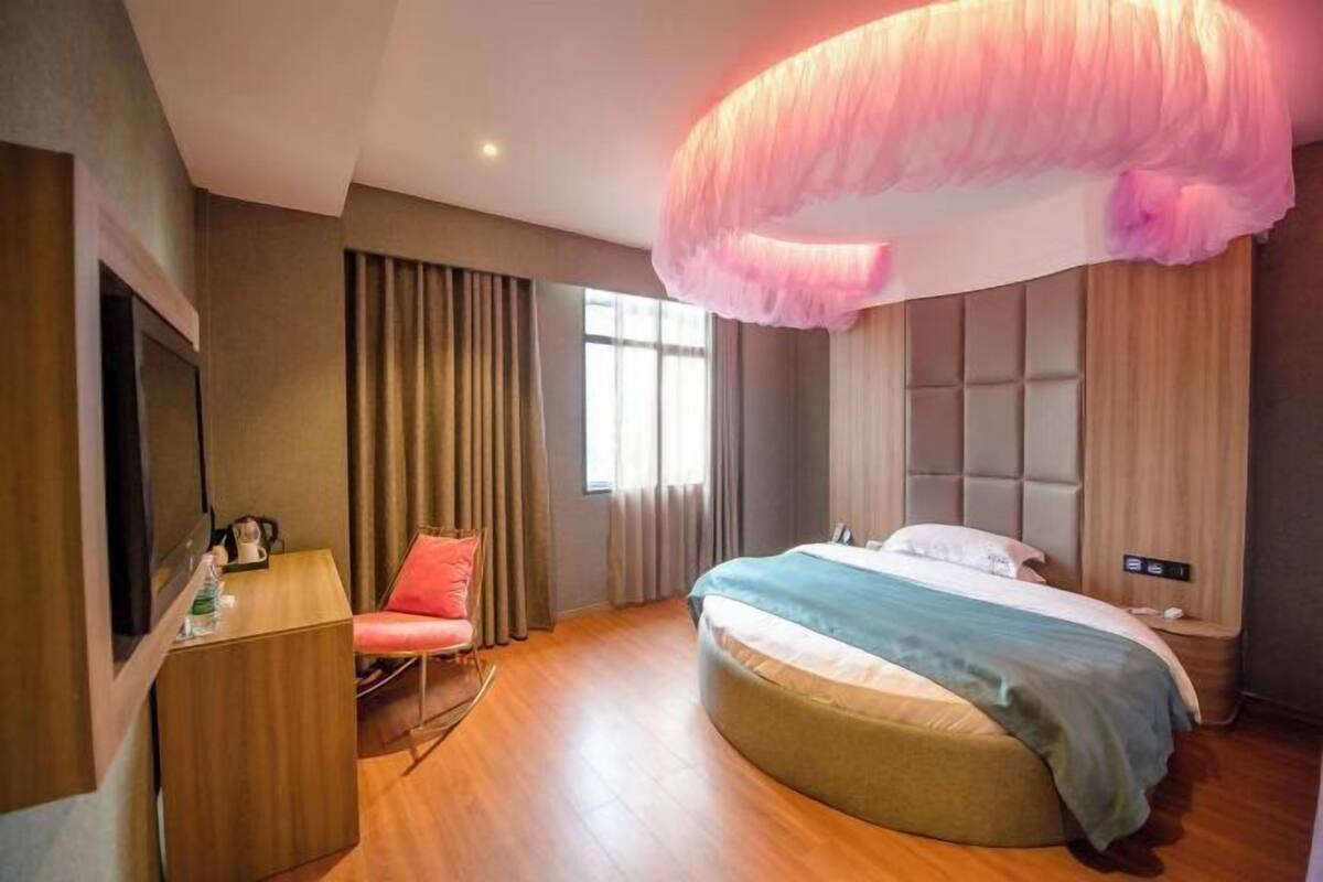 Une chambre d’hôtel moderne dotée d’un lit aux lignes courbes, d’un bureau avec une chaise, d’une télévision et d’une grande fenêtre avec des rideaux.