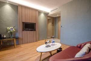 Una habitación de hotel con televisor de pantalla plana montado en la pared, una mesa de centro redonda con un florero, un sofá rojo y suelo de madera.