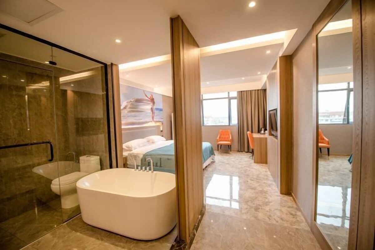 A modern hotel room with a large bathtub, a separate shower area, a bed, a seating area with a chair, and a large window offering a city view.