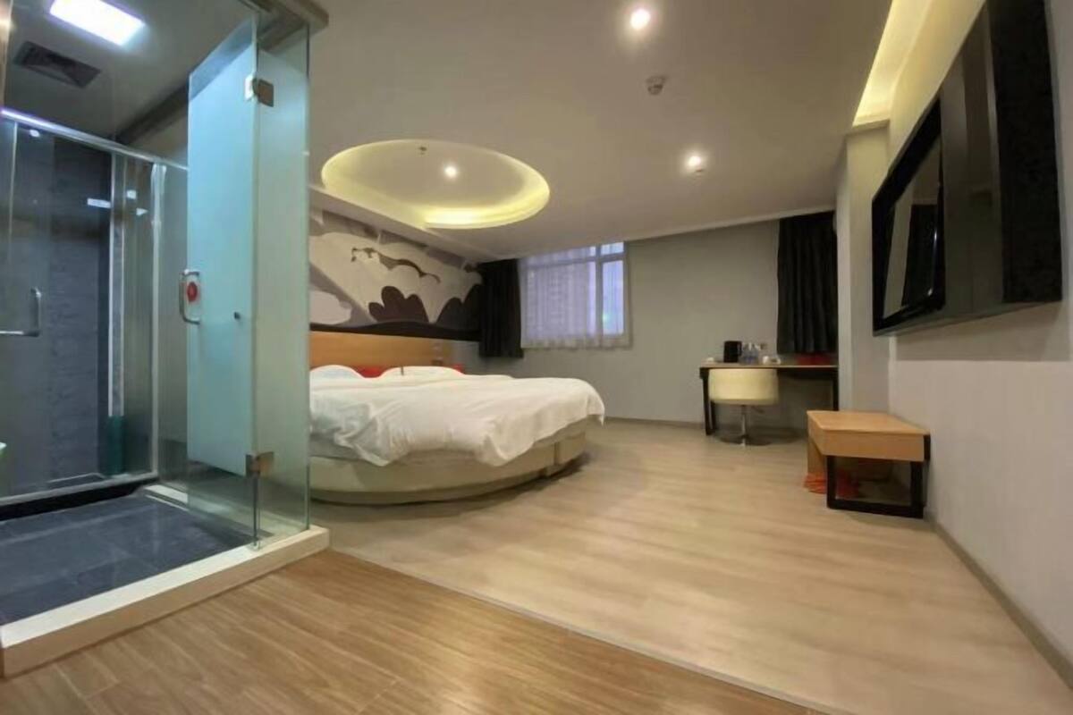 Une chambre d’hôtel moderne équipée d’un lit, d’un bureau, d’un miroir et d’une douche séparée par des parois en verre.