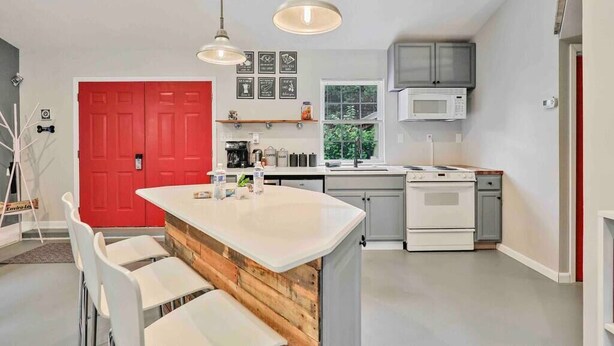 A modern kitchen with a central island, white cabinets, a red door, and a window with a view of greenery. - Fox Cottage *Pet Friendly* (Catonsville)
