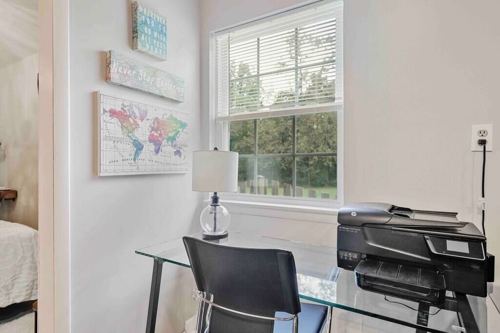 A room with a glass table, a black chair, a printer, a lamp, and a window with blinds.