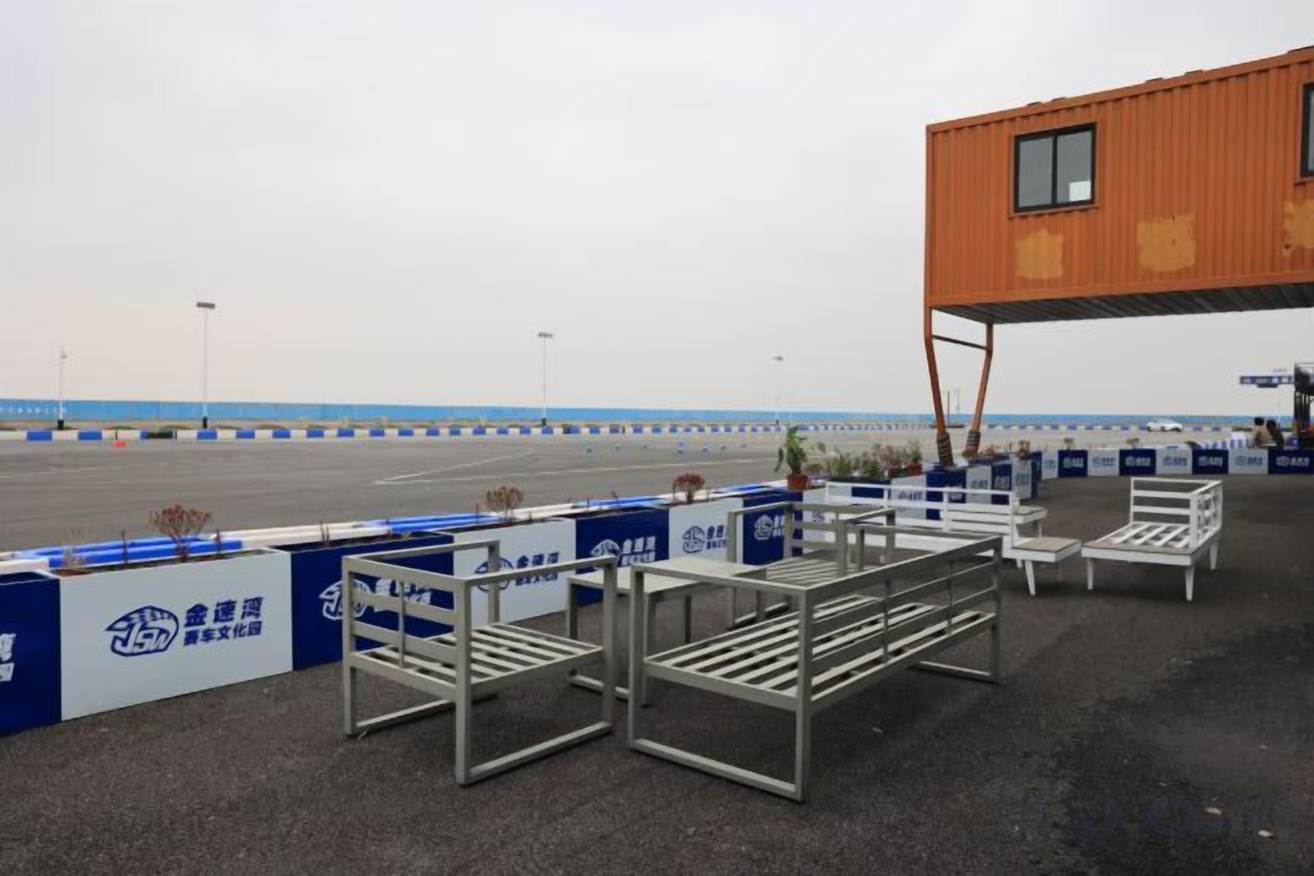 An outdoor seating area with metal tables and chairs, a building with a window, and a blue and white fence.