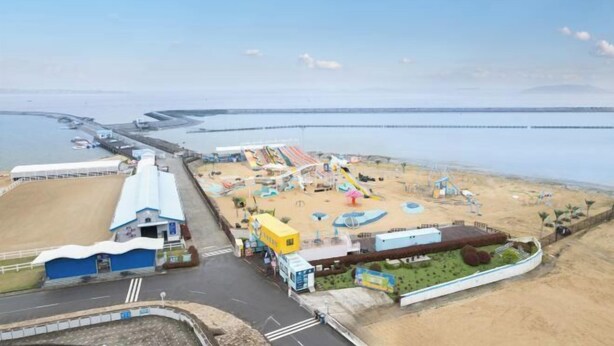 Luftaufnahme eines Küstenvergnügungsparks mit Sandstrand, Fahrgeschäften und Parkplatz.