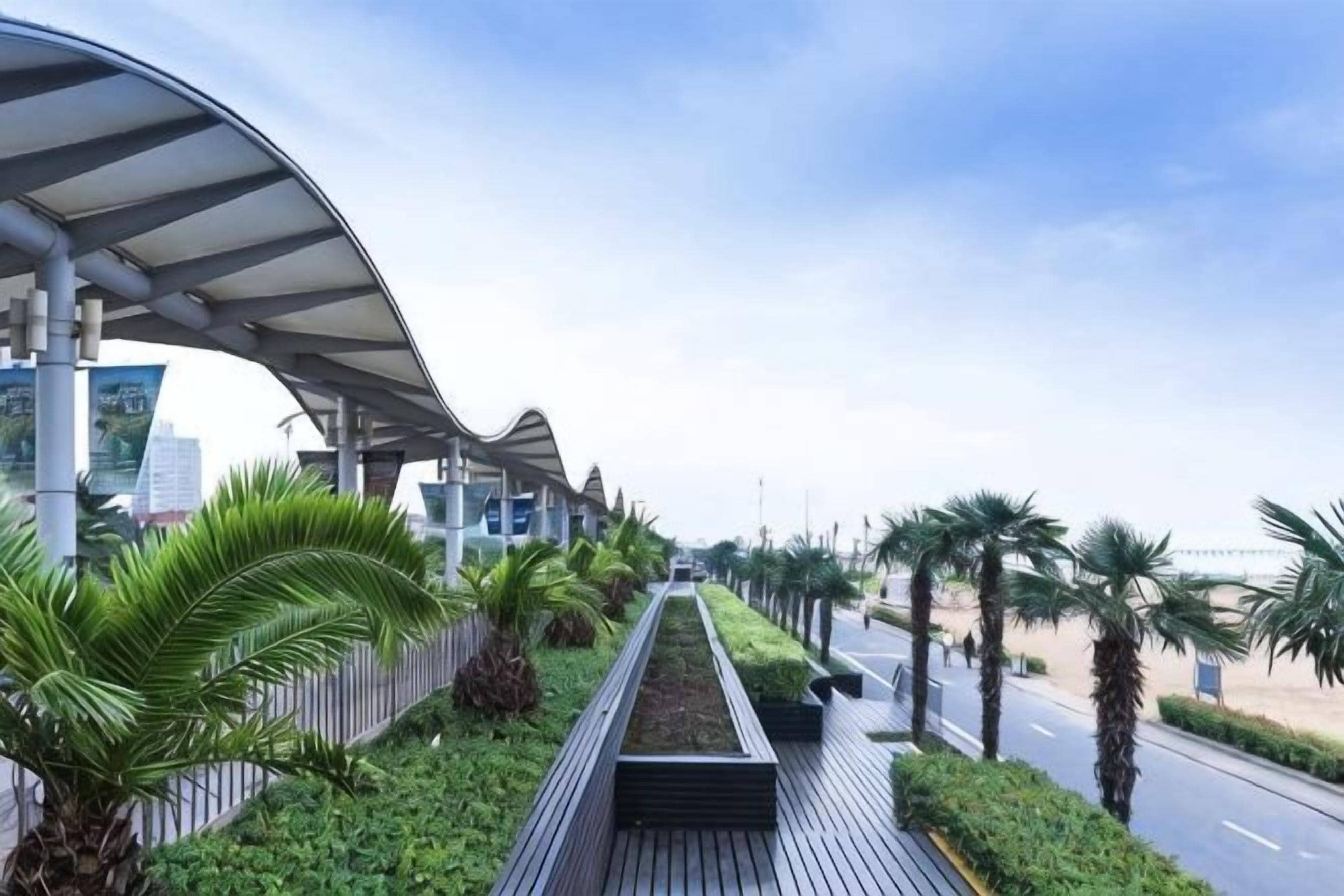 A covered walkway with a curved roof, lined with palm trees and greenery, leading to a road.